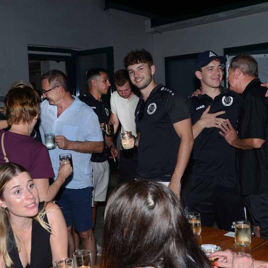 Eine Gruppe von Menschen, darunter mehrere Männer in schwarzen Uniformen, versammelt sich in einem Raum. Einige halten Gläser mit Getränken. Eine Frau lächelt in die Kamera. Der Raum hat weiße Wände und eine offene Tür.