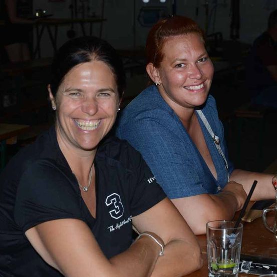 Zwei Frauen lächeln und sitzen zusammen an einem Tisch, eine trägt ein schwarzes Shirt mit der Nummer drei und die andere ein blaues Shirt. Ein Glas mit Strohhalm steht auf dem Tisch.