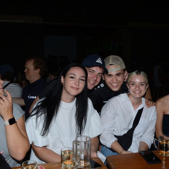 Eine Gruppe von Freunden lächelt in die Kamera. Zwei Frauen und zwei Männer posieren in einer Bar. Es gibt Getränke auf dem Tisch vor ihnen.