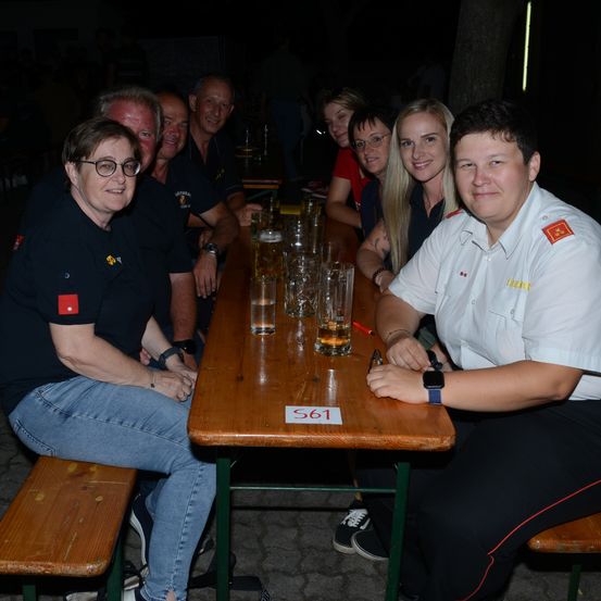 Eine Gruppe von Menschen sitzt an einem Tisch und lächelt für ein Foto. Sie tragen verschiedenfarbige Shirts, und der Tisch hat Gläser und die Nummer 561.