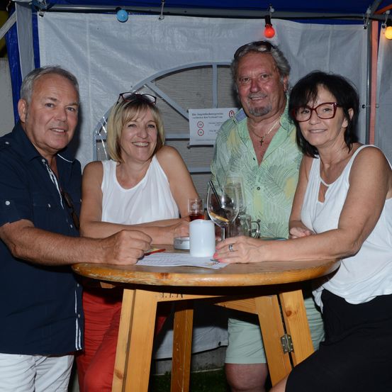 Vier Personen sitzen lächelnd an einem Tisch. Zwei Männer und zwei Frauen, eine Frau mit Brille und einem weißen Tanktop. Weingläser und Tassen auf dem Tisch. Hinter ihnen ein weißes Zelt mit einem Schild.