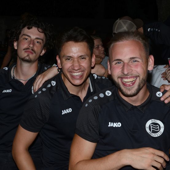Drei Männer tragen schwarze Jako-Trikots mit einem Logo auf der linken Brust und lächeln in die Kamera. Der Mann auf der rechten Seite hat einen Ring am Finger.