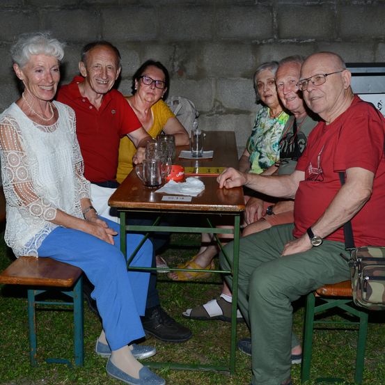 Eine Gruppe älterer Menschen sitzt lächelnd um einen Tisch. Zwei Frauen tragen Brillen und Halsketten, und die Männer tragen rote Hemden und Uhren. Leere Gläser und ein Teller stehen auf dem Tisch, und eine Wand ist im Hintergrund.