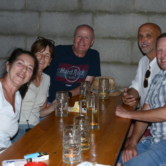 Fünf Menschen sitzen um einen Holztisch herum, lächeln und posieren für ein Foto. Auf dem Tisch stehen Gläser, ein Hut und etwas Essen. Der Mann in der Mitte trägt ein Hard Rock Cafe-Shirt.