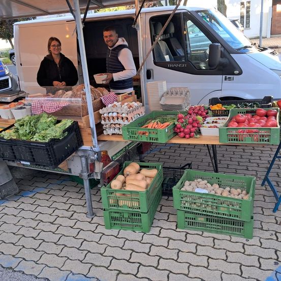 Zwei Personen stehen hinter einem Tisch voller frischer Produkte aus einem weißen Lieferwagen. Sie lächeln und halten Gegenstände in den Händen. Der Tisch steht auf einer Kopfsteinpflasterstraße.