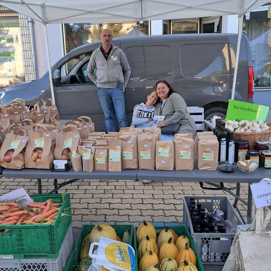 Zwei Frauen und ein junger Junge stehen hinter einem Tisch mit Tüten voller Obst und Gemüse. Hinter ihnen ist ein Van geparkt, und es gibt Kisten und Flaschen auf dem Tisch.
