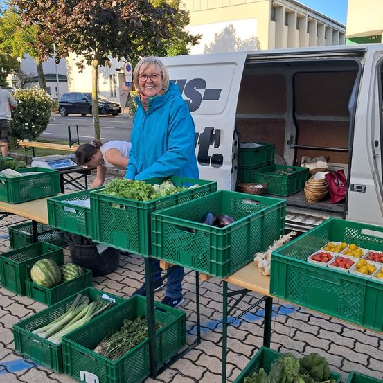 Eine Frau in einer blauen Jacke steht hinter einem Tisch voller grüner Kisten mit frischem Gemüse. Ein Lieferwagen mit den Buchstaben 'st' ist in der Nähe geparkt. Im Hintergrund befinden sich ein Baum, Gebäude und ein geparktes Auto.