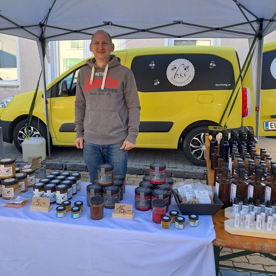 Ein Mann steht hinter einem Tisch mit verschiedenen Gläsern und Flaschen zur Schau. Er trägt eine graue Kapuzenjacke und Jeans. Hinter ihm steht ein gelber Van mit Bienenaufklebern.