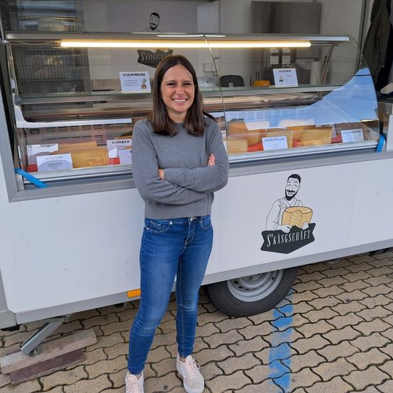 Eine Frau steht vor einem Lebensmittelwagen mit einer Vitrine voller Käseprodukte. Der Wagen hat ein Logo und den Text 'S'KASSGESCHAFT' an der Seite.