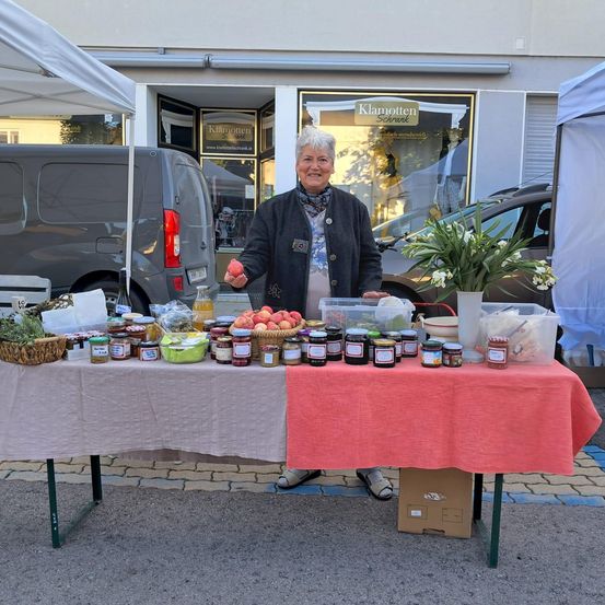 Eine Frau steht hinter einem Tisch mit verschiedenen Gläsern von Obstkonserven und einem Blumenstrauß. Sie ist draußen in der Nähe eines grauen Lieferwagens und eines Geschäfts.