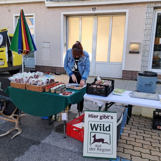 Eine Person steht an einem Tisch mit verschiedenen Lebensmitteln und Getränken. Ein bunter Regenschirm steht hinter dem Tisch. Ein Schild sagt 'Hier gibt's Wild aus der Region.' Es gibt einen gelben Van und ein Gebäude in der Nähe.