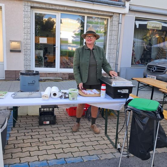 Eine Frau steht hinter einem Tisch, trägt eine Mütze und Brille und hält ein Kochgerät. Der Tisch hat verschiedene Gegenstände, darunter Papierrollen, Behälter und ein Schneidebrett. Im Hintergrund ist ein Geschäft mit Glastüren zu sehen.