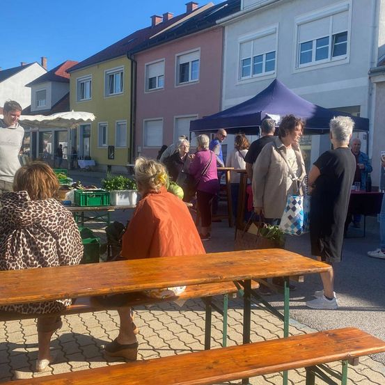 Eine Gruppe von Menschen versammelt sich um einen Holztisch auf einem Markt im Freien. Sie kaufen bei Ständen ein, und im Hintergrund ist ein Zelt aufgebaut.