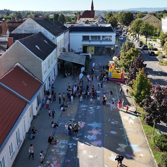Eine Luftaufnahme einer Straße mit Kreidezeichnungen, Menschen beim Gehen und Sitzen. Es gibt Bäume, Gebäude und eine Straße mit Autos. Ein Gebäude mit einem Turm ist in der Ferne sichtbar.