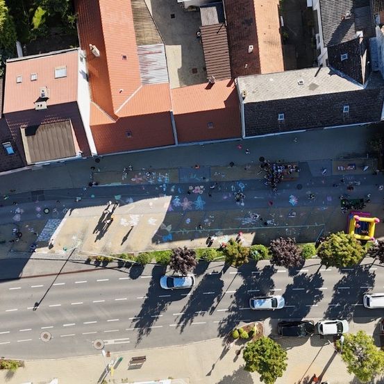 Luftaufnahme eines Wohngebiets mit Gebäuden, Menschen, Kreidezeichnungen, Autos und Bäumen. Einige Menschen gehen auf dem Bürgersteig.