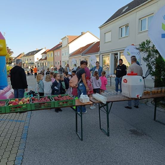 Eine Gruppe von Menschen versammelt sich um einen Tisch mit Äpfeln und Getränken auf einer Straße in einer Stadt. Im Hintergrund befinden sich einige Häuser und eine Fahne.
