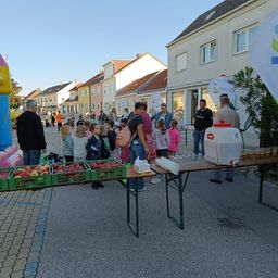 Eine Gruppe von Menschen versammelt sich um einen Tisch mit Äpfeln und Getränken auf einer Straße in einer Stadt. Im Hintergrund befinden sich einige Häuser und eine Fahne.