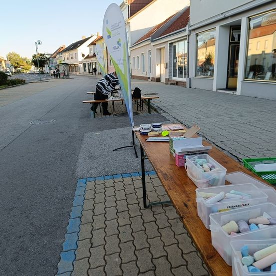Eine leere Straße mit einem langen Holztisch voller Kreide auf einem Bürgersteig. Eine Person sitzt auf einer Bank in der Nähe.