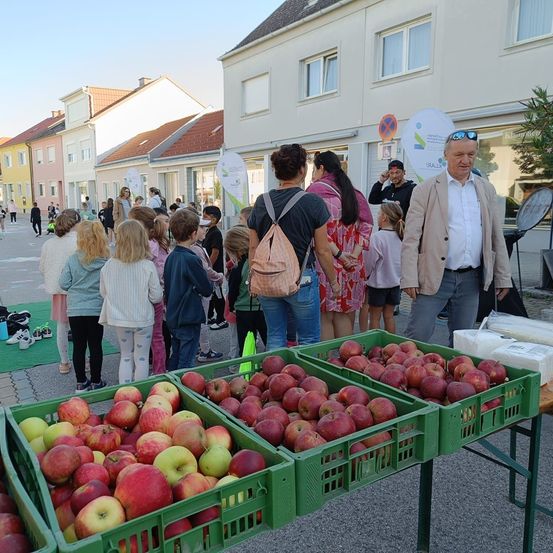 Menschen versammeln sich um einen Tisch mit roten und grünen Äpfeln, wobei mehrere Personen Brillen und Rucksäcke tragen. Ein Mann in einem hellen Jackett steht neben dem Tisch. Dahinter befinden sich Häuser und Gebäude, mit einem klaren Himmel darüber.