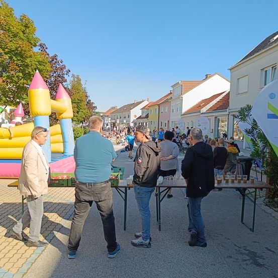 Menschen versammeln sich um einen Tisch mit Getränken im Freien an einem sonnigen Tag, in der Nähe eines Springkastens und Gebäuden.