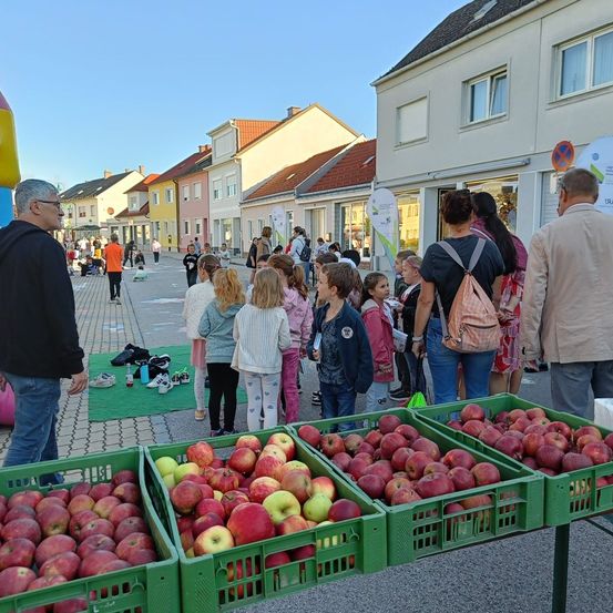 Ein Outdoor-Markt mit verschiedenen Menschen, einschließlich Kindern, die Körbe mit roten Äpfeln auf einem Tisch vor Gebäuden betrachten.