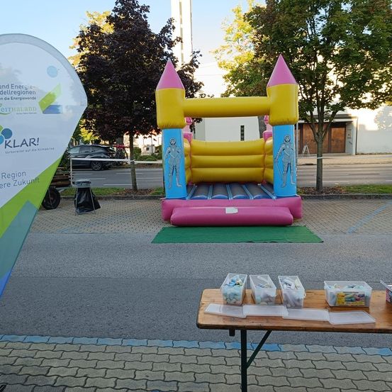 Ein aufblasbarer Burg steht auf einem Parkplatz, davor ein Tisch mit verschiedenen Spielzeugen. Bäume und ein weißes Gebäude sind im Hintergrund zu sehen.