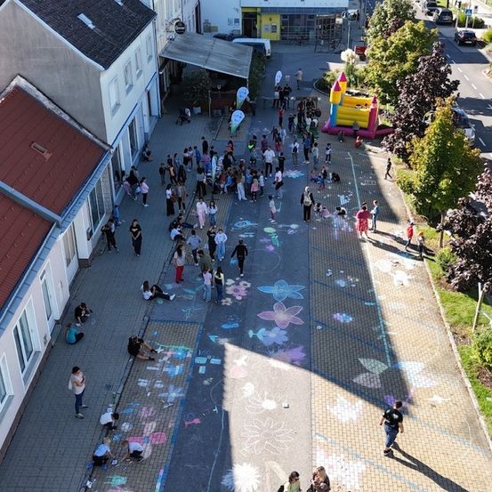 Eine Vogelperspektive einer Straße mit vielen Menschen, einige malen auf der Straße, andere gehen oder sitzen. Ein Hüpfburg und ein Zelt sind aufgestellt.