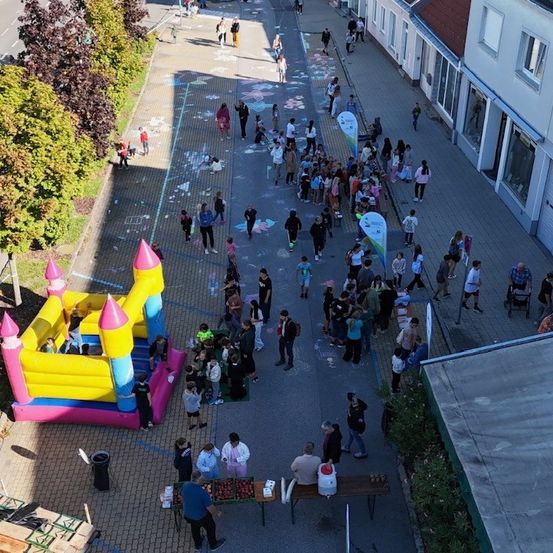 Luftaufnahme einer geschäftigen Straßenveranstaltung mit Menschen und einem aufblasbaren Schloss. Einige gehen, andere stehen herum und einige sitzen auf Bänken. Bunte Kreidezeichnungen schmücken den Boden.