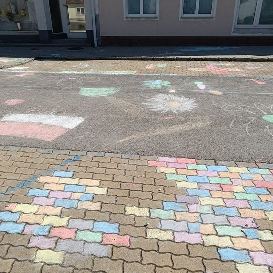 Ein Gehweg ist mit bunten Kreidezeichnungen, einschließlich Blumen und geometrischer Formen, vor einem Gebäude mit Glasfenstern verziert.