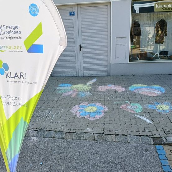 Ein Banner mit dem Wort 'KLAR!' steht vor einem Gebäude mit Schaufenster. Kreidezeichnungen von Blumen sind auf dem Boden.