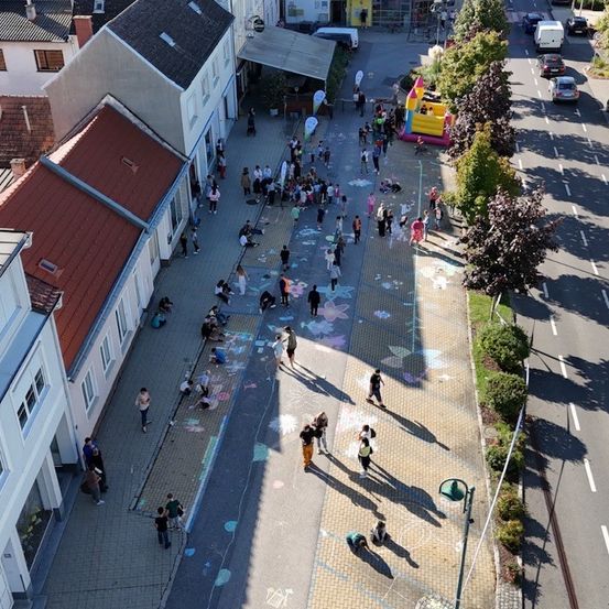 Luftaufnahme einer belebten Straße mit Kreidezeichnungen auf dem Bürgersteig, spielenden Kindern und einem Hüpfburg. Gebäude säumen die Straße, mit einem klaren Blick auf die Straße und geparkte Autos.