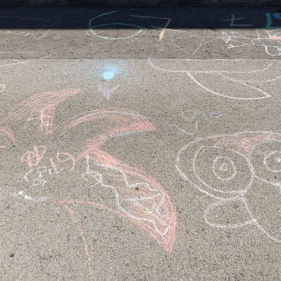 Ein Betonpflaster mit verschiedenen Kreidezeichnungen, darunter ein Fisch, ein Gesicht und andere Formen, einige in Rosa und Blau. Schatten fallen auf das Pflaster.