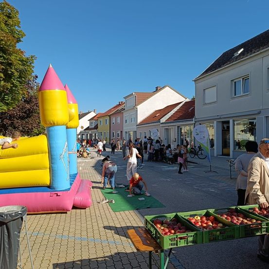 Eine lebhafte Straßenszene mit einem farbenfrohen aufblasbaren Schloss, gehenden Menschen und einem Obststand. Der Bereich ist voller Aktivität, mit einer Gruppe von Menschen, die in der Nähe von Gebäuden stehen, und einem Fahrrad, das in der Nähe geparkt ist.