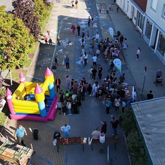 Eine Luftaufnahme einer belebten Straße mit vielen Menschen, die gehen, einige spielen auf einem Bürgersteig mit Kreidezeichnungen, und ein Hüpfburg steht links. Eine Gruppe von Menschen versammelt sich um einen Tisch mit Essen. Bäume und Pflanzen befinden sich an den Straßenrändern.