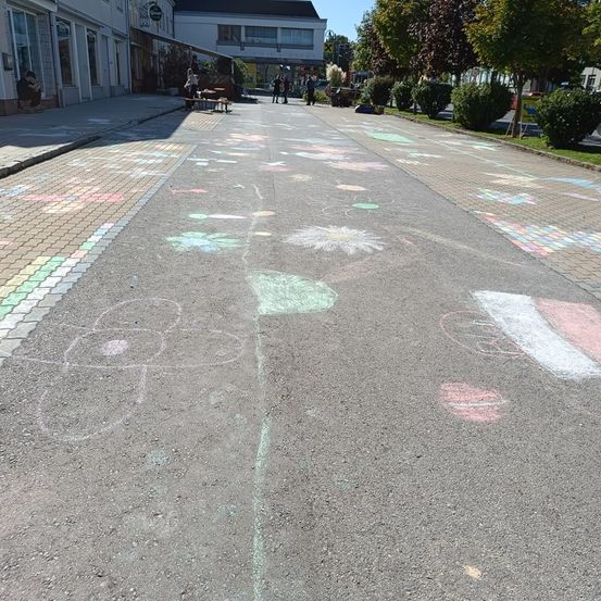Ein Gehweg mit bunten Kreidezeichnungen, darunter Blumen und Herzen, erstreckt sich in eine Straße mit Ziegelpflaster. Im Hintergrund sind Menschen in der Nähe von Gebäuden und Grünflächen zu sehen.