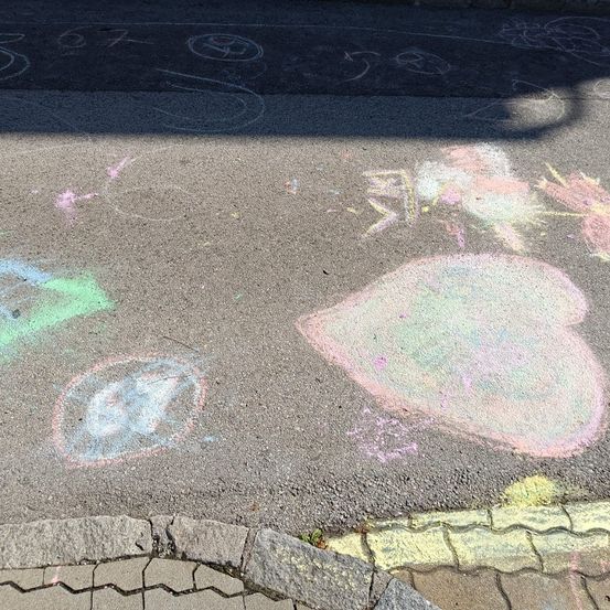 Ein Bürgersteig mit verschiedenen farbigen Kreidezeichnungen, darunter ein großes Herz, ein Kreis und einige abstrakte Formen. Der Bürgersteig hat eine Ziegelsteingrenze.