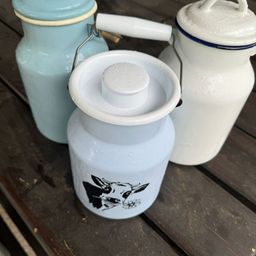 Drei Milchkrüge stehen auf einem Holztisch. Der mittlere Krug hat eine Kuh-Illustration, während die anderen beiden schlicht sind.