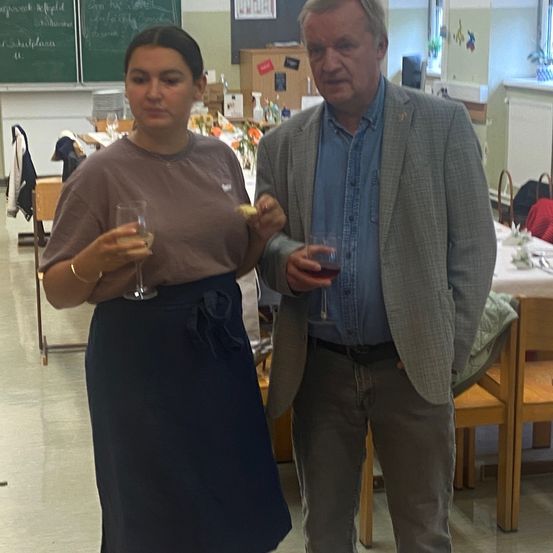 Ein Mann und eine Frau stehen zusammen, Weinglas in der Hand, in einem, was einem Speisesaal zu ähneln scheint, mit Stühlen, einem Tisch und einer Tafel.