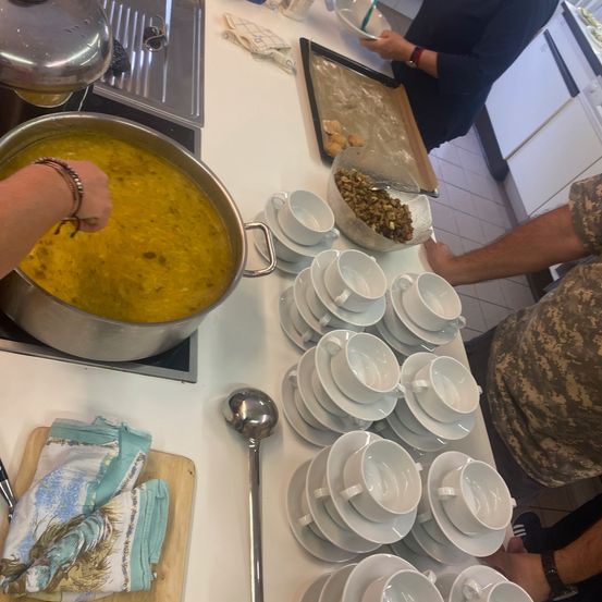 Eine Gruppe von Menschen kocht in einer Küche. Ein großer Topf mit Suppe steht auf dem Herd, während eine Person einen Schöpflöffel hält. Verschiedene Gerichte, Schüsseln und Utensilien sind auf dem Tresen angeordnet. Ein Tablett mit Essen und eine Schüssel mit mehr Essen stehen auf dem Tisch.