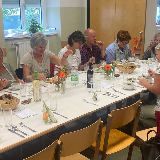 Eine Gruppe Erwachsener sitzt um einen Tisch mit Essen, Wein und Blumen. Der Tisch ist mit Tellern, Gläsern und Besteck gedeckt. Eine Frau in einer weißen Schürze lächelt.