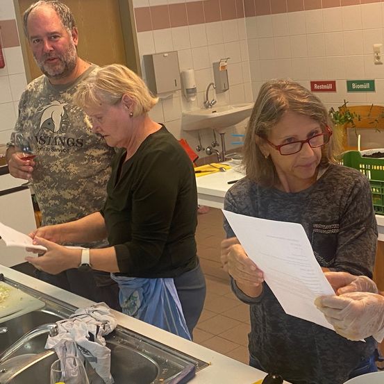 Vier Personen stehen in einer Küche und halten Papiere. Eine Frau hält ein Weinglas. Eine andere Frau hält ein Papier, während eine andere es betrachtet. Ein Waschbecken und eine Theke stehen vor ihnen.