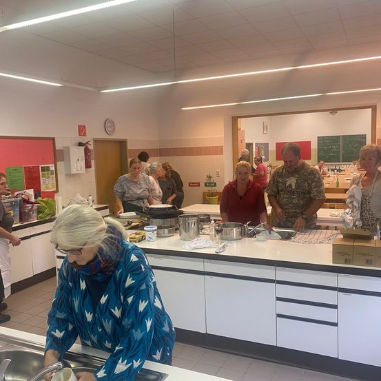 Mehrere Menschen versammeln sich in einer Küche, wobei eine Frau Geschirr spült. Ein Tisch ist mit Töpfen, Pfannen und einer Schüssel beladen, während andere Gespräche führen. Die Küche verfügt über einen Feuerlöscher, Schränke und eine Uhr an der Wand.