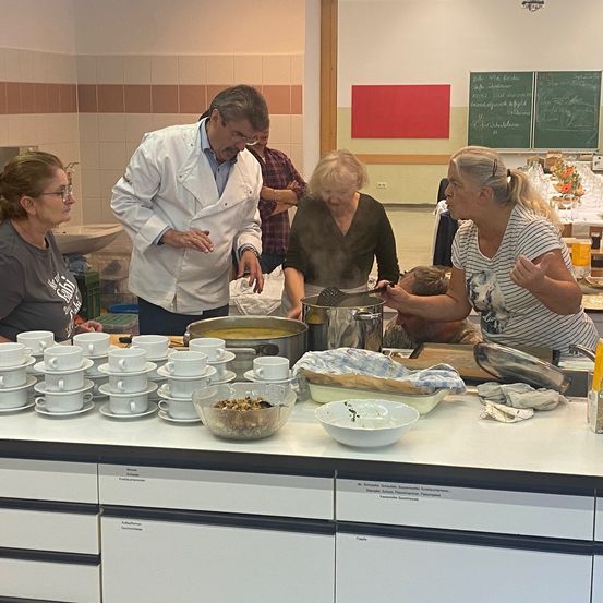 Eine Gruppe von Menschen in einer Küche, ein Mann in einer Kochjacke erklärt etwas, andere kochen. Tassen, Schüsseln und Töpfe auf der Arbeitsplatte. Ein Tafel im Hintergrund.