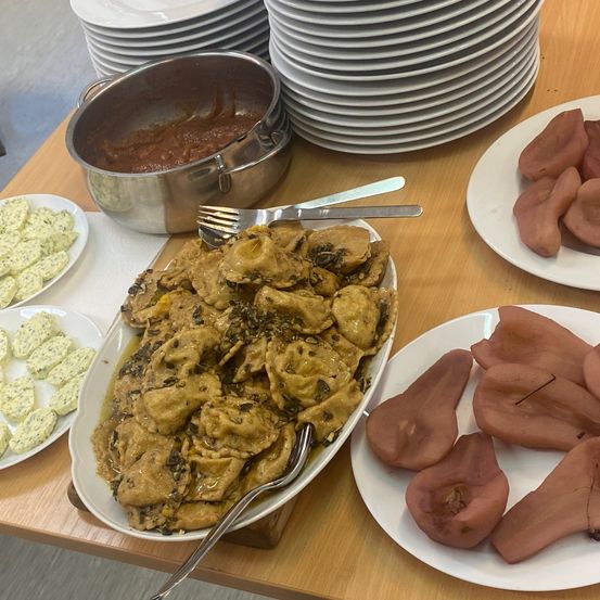 Ein Holztisch hat verschiedene Gerichte darauf. Ein Teller mit einem Fleischgericht, ein anderer mit Birnenscheiben und ein Teller mit einem Käsesnack. Besteck, gestapelte Teller und ein Topf mit Essen sind ebenfalls auf dem Tisch.