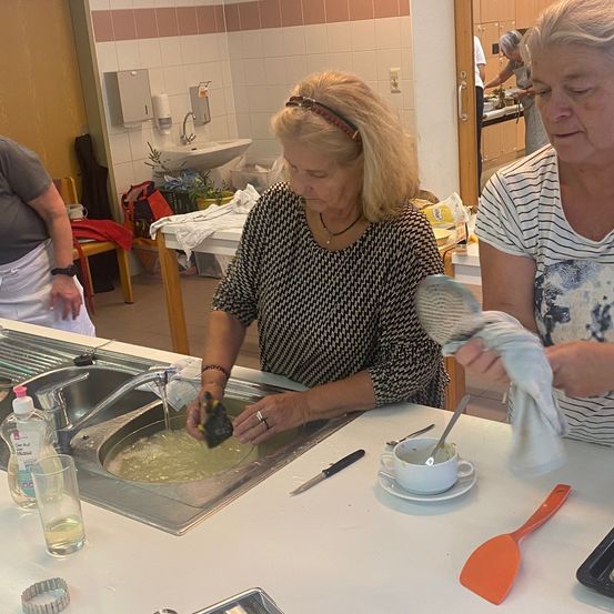 Drei Frauen sind in einer Küche. Eine wäscht Geschirr, eine andere hält ein Handtuch und eine dritte steht mit den Händen auf den Hüften. Ein Waschbecken, Geschirr und Küchenutensilien sind sichtbar.