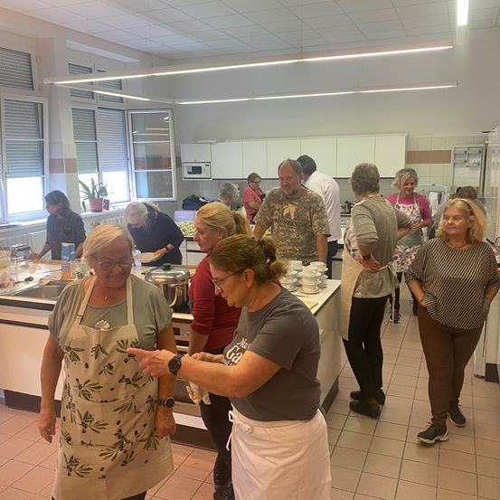 Eine Gruppe von Menschen, einige in Schürzen, versammelt sich in einer Küche. Sie stehen und unterhalten sich. Einige bereiten Essen zu, während andere beobachten. Der Raum ist gut beleuchtet und mit modernen Geräten ausgestattet.