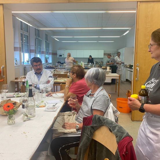 Mehrere Personen sind in einer Küche versammelt. Ein Mann in einem weißen Laborkittel sitzt an einem Tisch mit anderen. Sie haben Flaschen, Weingläser und einen Korb mit Blumen. Ein Lehrer steht in der Nähe.