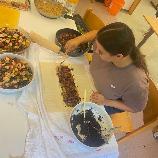 Eine Frau bereitet ein Gericht auf einem Tisch zu, umgeben von verschiedenen Zutaten und Utensilien. Sie verteilt eine Mischung auf ein Teigblatt. Schüsseln mit Obst und Gemüse, ein Nudelholz und ein Messer stehen auf dem Tisch.