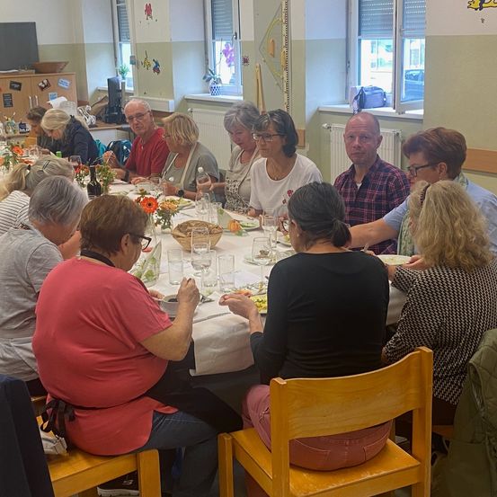 Eine Gruppe älterer Menschen sitzt um einen langen Tisch herum und genießt eine Mahlzeit. Sie sitzen auf Holzstühlen, und der Tisch ist mit einer weißen Tischdecke bedeckt. Blumen und Kürbisse sind auf dem Tisch platziert, und im Hintergrund befinden sich Fenster mit weißen Jalousien.