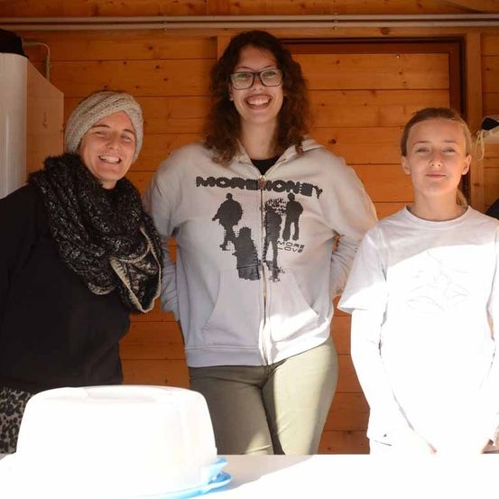 Drei Frauen stehen in einer Hütte mit Holzwänden. Ein Kuchen steht auf dem Tisch. Die Frau links trägt eine Mütze und einen Schal. Die Frau in der Mitte trägt eine Brille und eine Kapuzenjacke. Die Frau rechts trägt ein weißes T-Shirt.